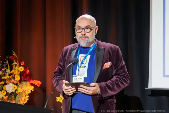 Mężczyzna w okularach, w eleganckim garniturze, przemawia na scenie z tabletem w ręku.