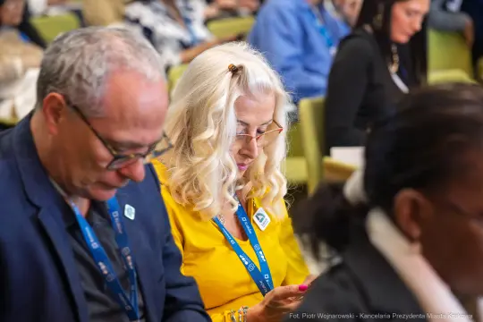 Kobieta z blond włosami w żółtej bluzce korzysta z telefonu, siedząc wśród innych uczestników konferencji.