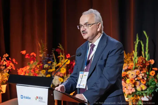 Mężczyzna przemawia z mównicy podczas konferencji, w tle kolorowe kwiaty.