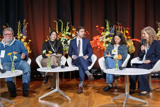 Panel dyskusyjny z pięcioma uczestnikami siedzącymi przy okrągłych stołach, w tle kwiaty.