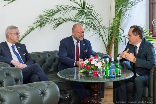 Spotkanie trzech mężczyzn w eleganckim wnętrzu z kwiatami i napojami na stole.