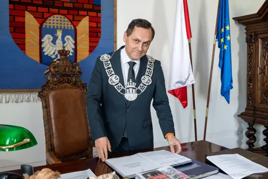 Mężczyzna w garniturze z łańcuchem na szyi stoi przy biurku w otoczeniu flag i herbów.
