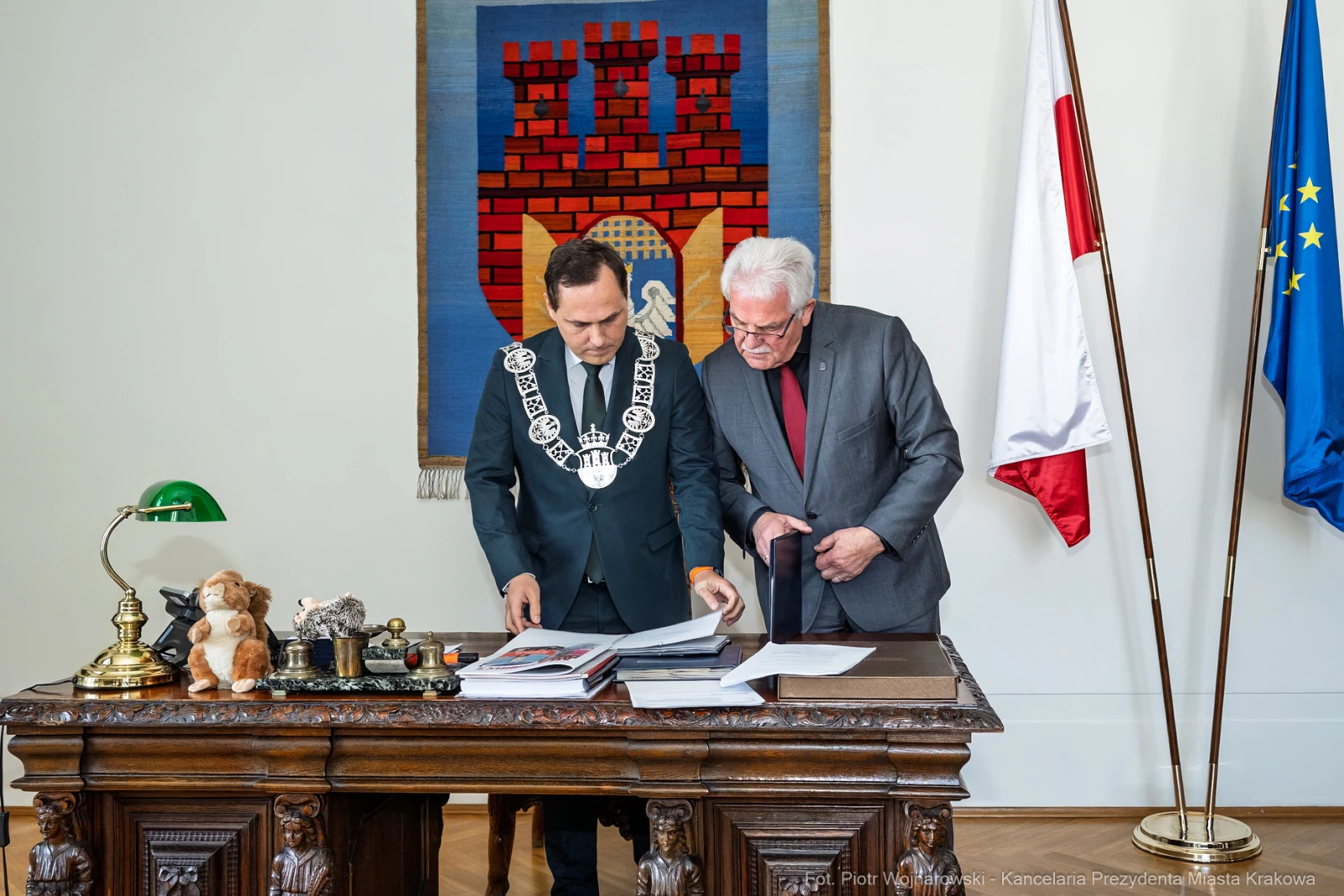 Dwaj mężczyźni przy biurku, przeglądają dokumenty, w tle flagi i herb miasta.  Autor: Piotr Wojnarowski