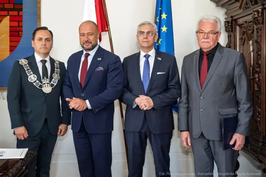 Czterech mężczyzn w garniturach, stojących obok siebie, w tle flagi Polski i Unii Europejskiej.