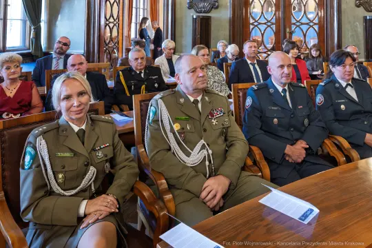 Grupa osób w mundurach i garniturach siedząca w sali podczas uroczystości.
