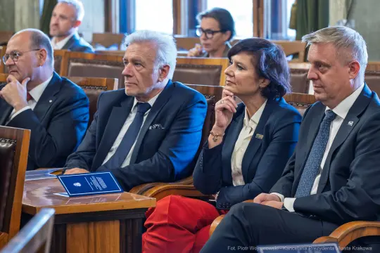 Cztery osoby siedzące w sali, skupione na wystąpieniu, w formalnych strojach.