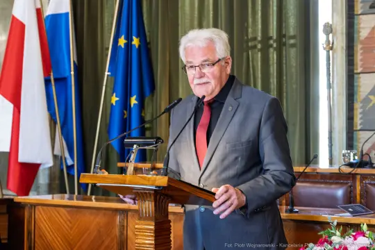 Mężczyzna w garniturze przemawia z mównicy, w tle flagi Polski, Unii Europejskiej i innych.