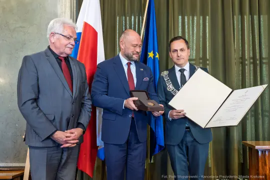 Wręczenie medalu i dyplomu w uroczystej atmosferze, trzech mężczyzn w garniturach.