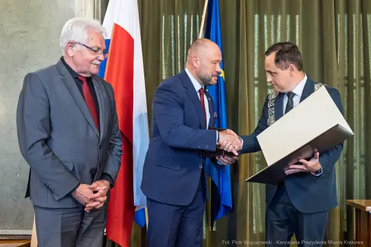 Wręczenie nagrody w uroczystej atmosferze, trzech mężczyzn w formalnych strojach.
