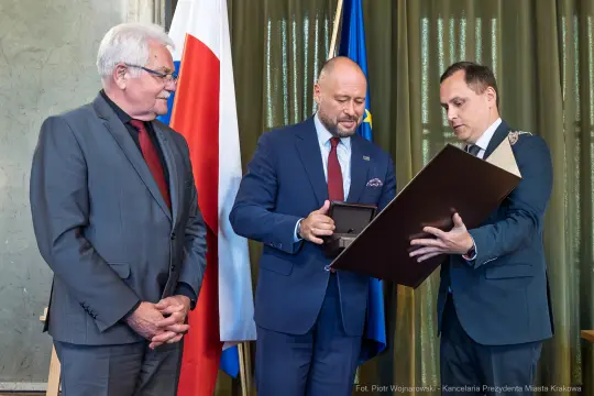 Trzech mężczyzn w garniturach, wręczanie odznaczenia w obecności flagi Polski i Unii Europejskiej.