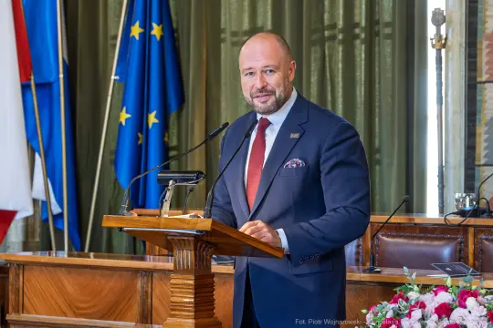 Mężczyzna w garniturze przemawia przy mównicy, w tle flagi Polski i Unii Europejskiej.