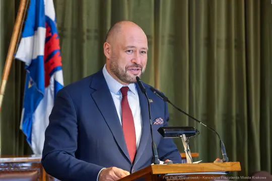 Mężczyzna w garniturze przemawia przy mównicy, w tle flagi i zasłony.