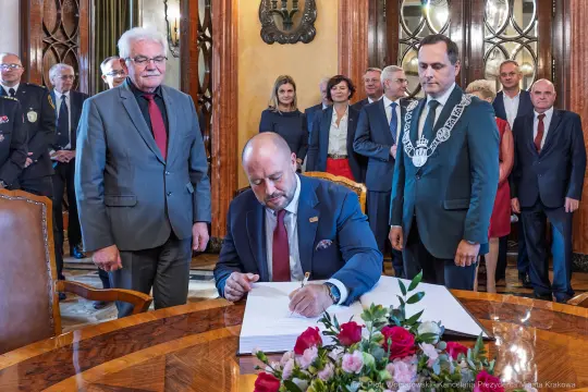 Mężczyzna podpisuje dokument w otoczeniu innych osób w eleganckim wnętrzu.