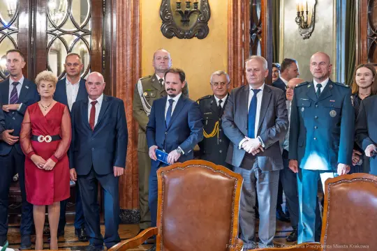 Grupa ludzi w eleganckich strojach w sali z dekoracjami, w tle widoczne oświetlenie i meble.