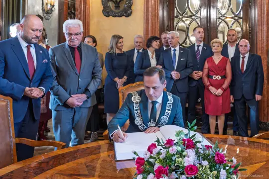 Mężczyzna w łańcuchu prezydenckim podpisuje dokument w otoczeniu gości. Kwiaty na stole.
