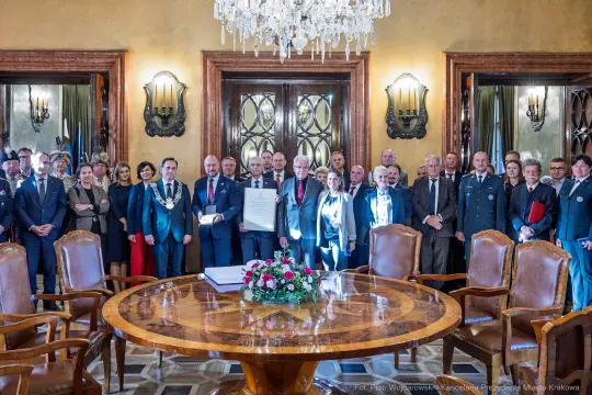 Grupa ludzi w formalnych strojach, stojących wokół okrągłego stołu w eleganckim wnętrzu.