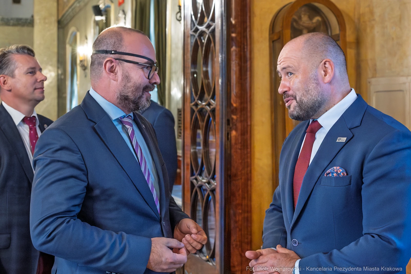 Dwaj mężczyźni rozmawiają w eleganckim wnętrzu, jeden z nich w garniturze z przypinką.  Autor: Piotr Wojnarowski