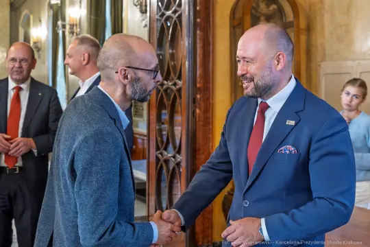 Spotkanie dwóch mężczyzn w formalnych strojach, uściski dłoni, w tle inni uczestnicy.