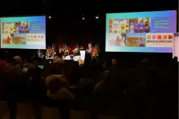 Konferencja z prezentacjami, uczestnicy siedzą na widowni, kwiaty dekoracyjne w tle.