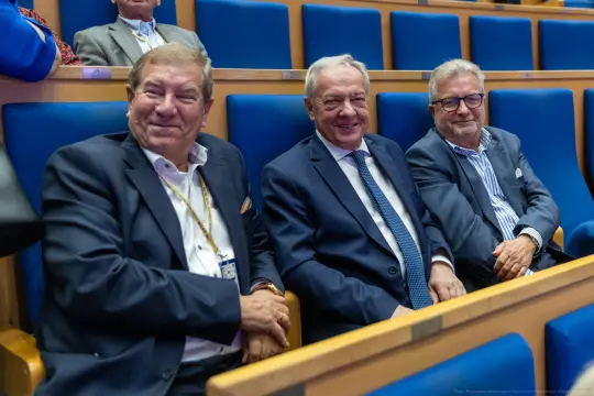 Trzech mężczyzn w garniturach siedzących na niebieskich fotelach w sali konferencyjnej.