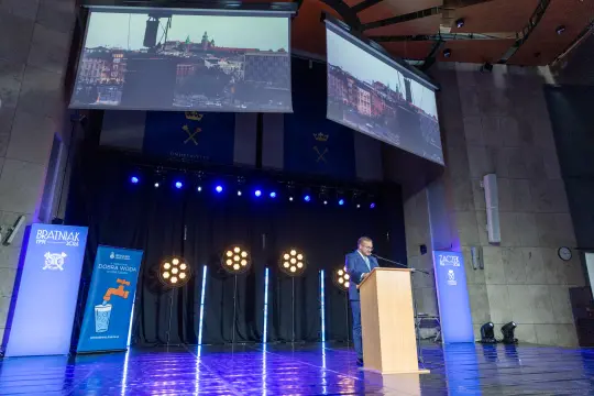 Mężczyzna przemawia z podium na tle oświetlonej sceny i ekranów wyświetlających widok miasta.