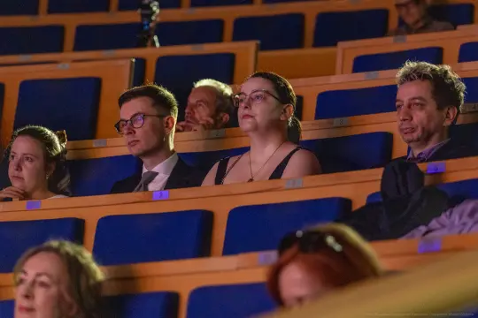Grupa ludzi siedzących na widowni, skupionych na wydarzeniu.