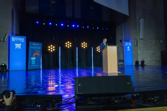 Mężczyzna przemawia z podium na tle oświetlonej sceny podczas wydarzenia.