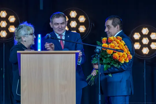Trzech mówców na scenie, jeden z bukietem pomarańczowych róż.