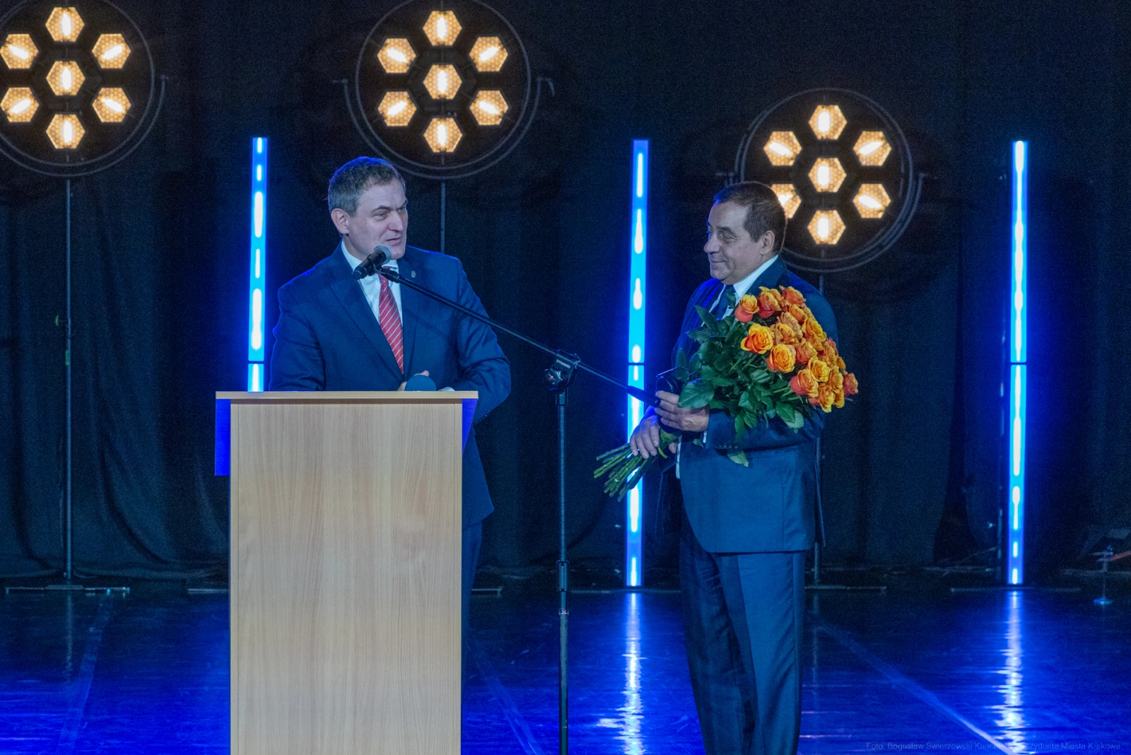 Dwaj mężczyźni na scenie, jeden z bukietem pomarańczowych róż, drugi przy mikrofonie.  Autor: Bogusław Świerzowski