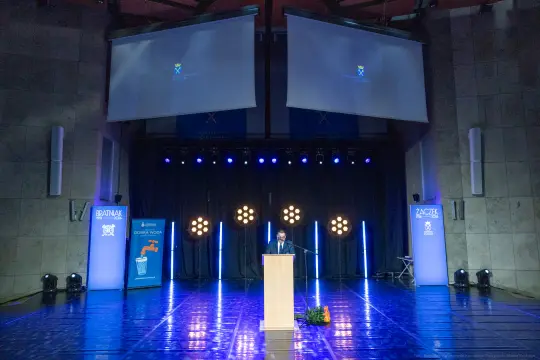 Mężczyzna przemawia z podium na scenie oświetlonej reflektorami. W tle flagi i plakaty.