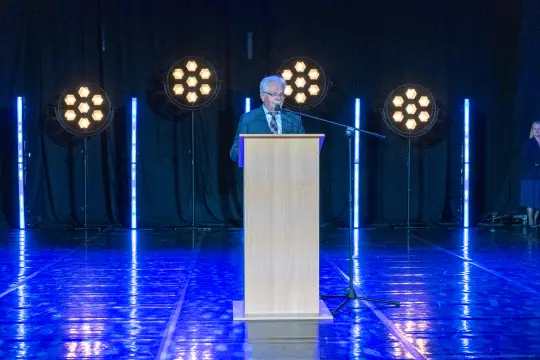 Mężczyzna przemawia z podium, w tle oświetlenie sceniczne.
