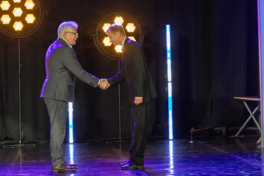 Dwaj mężczyźni w garniturach podają sobie ręce na scenie.