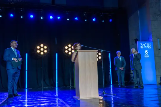Kobieta przemawia z podium, w tle mężczyźni i oświetlenie sceniczne.
