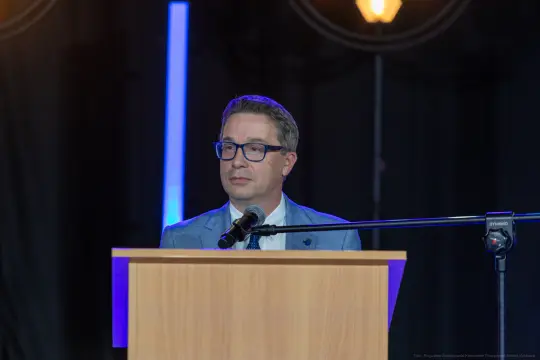Mężczyzna w okularach stoi przy mównicy, wygłasza przemówienie.