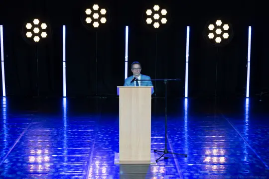 Mężczyzna przemawia z podium na tle oświetlenia scenicznego.