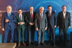 Grupa mężczyzn w garniturach na tle niebieskiej ściany z logo i napisami.