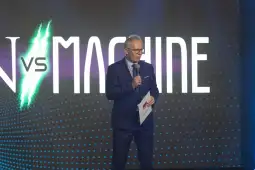 Mężczyzna w garniturze trzyma mikrofon, stoi przed dużym napisem "MAN vs MACHINE".