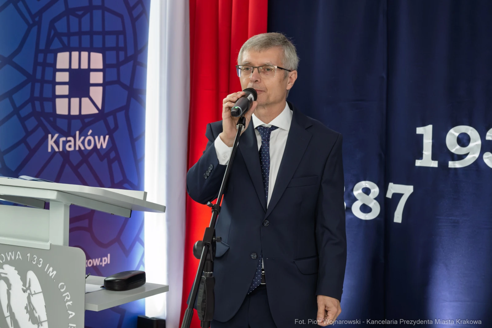 Mężczyzna w garniturze przemawia do mikrofonu na tle flagi i dekoracji.  Autor: Piotr Wojnarowski