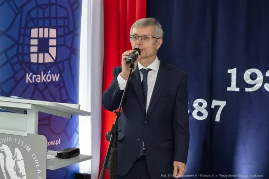 Mężczyzna w garniturze przemawia do mikrofonu na tle flagi i dekoracji.