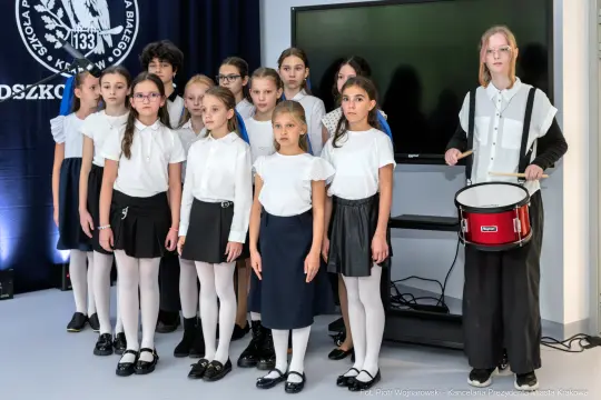 Grupa dzieci w strojach galowych, stojących na scenie, z perkusistą z boku.