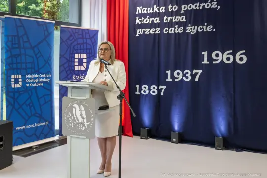 Kobieta przemawia na tle banerów Miejskiego Centrum Obsługi Oświaty w Krakowie.