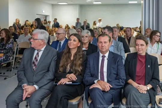 Grupa ludzi siedzących na sali podczas wydarzenia, w tle widoczni uczestnicy.