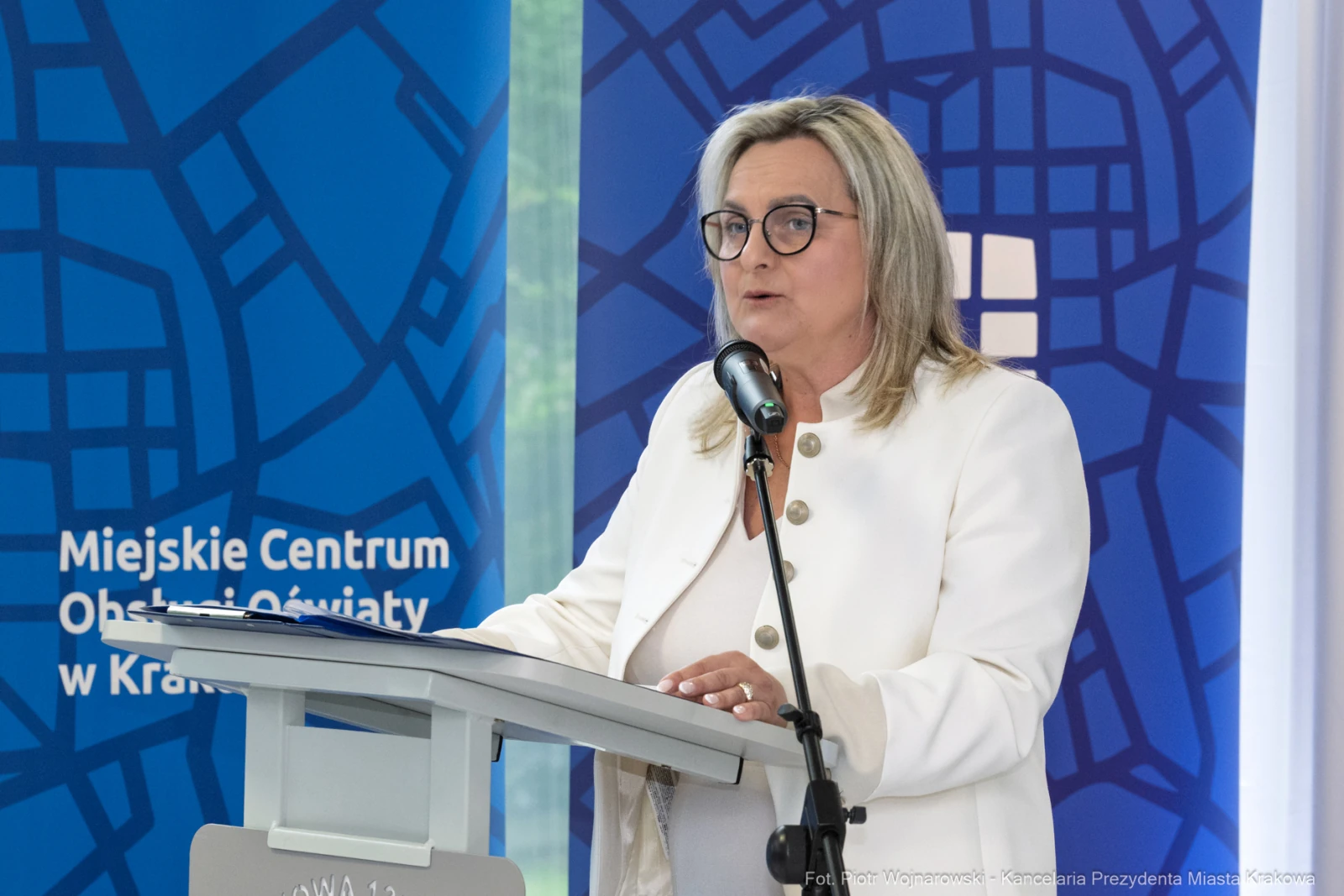 Kobieta przemawia przy mównicy, w tle niebieskie tło z logo Miejskiego Centrum Obsługi Oświaty w Krakowie.  Autor: Piotr Wojnarowski