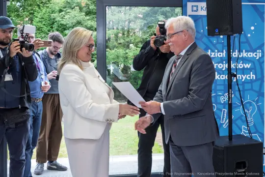 Wręczenie dyplomu w obecności mediów, uśmiechnięte osoby w formalnych strojach.