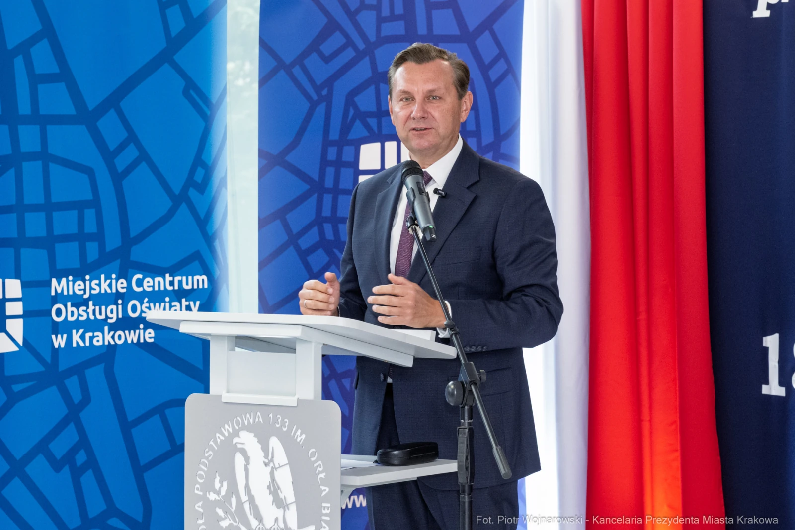 Mężczyzna przemawia przy mównicy, w tle flagi i logo Miejskiego Centrum Obsługi Oświaty w Krakowie.  Autor: Piotr Wojnarowski