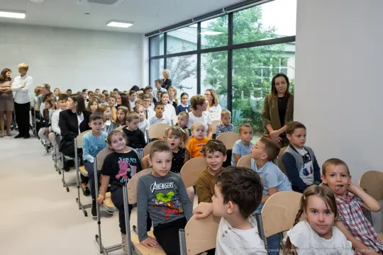 Grupa dzieci siedzących w sali, uśmiechających się i rozmawiających.