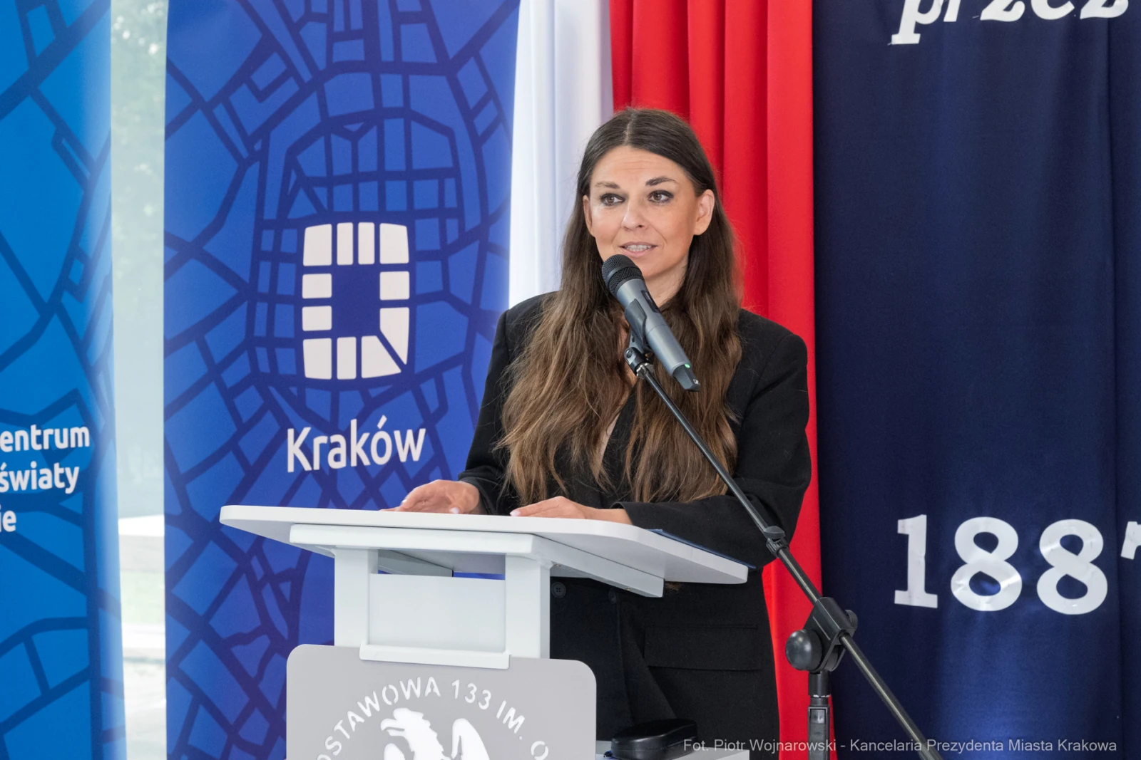 Kobieta przemawia przy mównicy, w tle flagi i logo Krakowa.  Autor: Piotr Wojnarowski