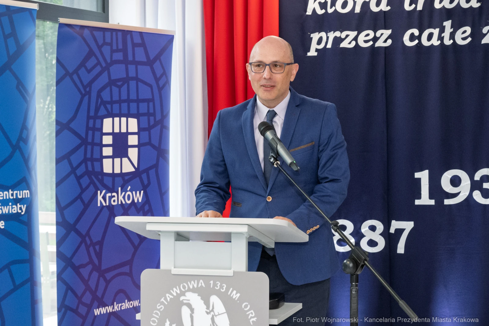 Mężczyzna w garniturze przemawia przy mównicy, w tle flagi i banery z logo Krakowa.  Autor: Piotr Wojnarowski