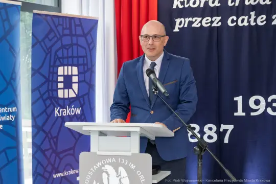 Mężczyzna w garniturze przemawia przy mównicy, w tle flagi i banery z logo Krakowa.