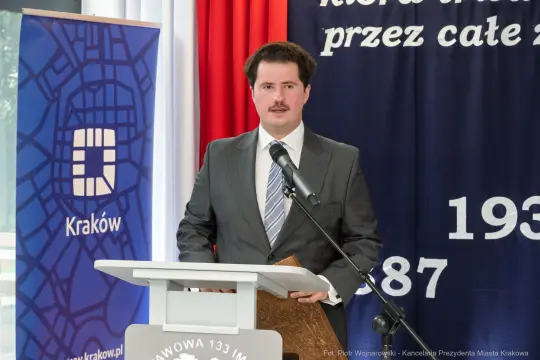 Mężczyzna w garniturze przemawia przy mównicy, w tle flagi i baner z logo Krakowa.